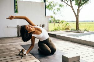 A woman doing yoga stretches on a porch with a kitten beside her in a serene outdoor setting.